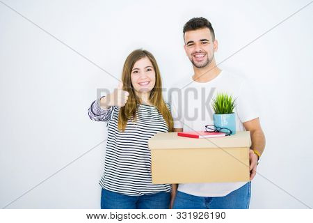 Young couple holding cardboard box moving to a new home over white isolated background happy with big smile doing ok sign, thumb up with fingers, excellent sign