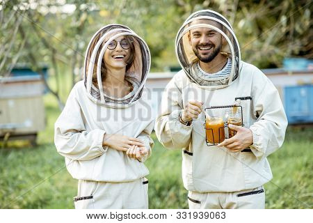 Portrait Of A Man And Woman Beekepers In Protective Uniform Standing Together With Honey In The Jar,