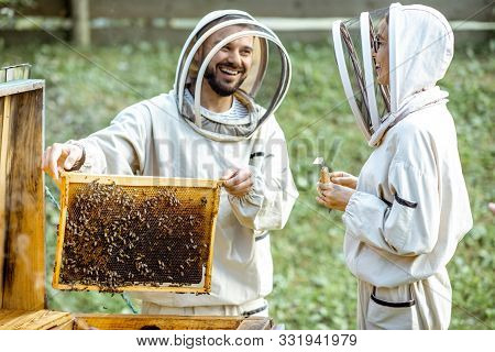Two Young Beekepers In Protective Uniform Working On A Small Apiary Farm, Getting Honeycomb From The