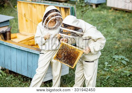Two Young Beekepers In Protective Uniform Working On A Small Apiary Farm, Getting Honeycombs From Th
