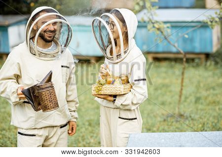 Two Beekepers In Protective Uniform Standing Together With Beesmoker And Honey, Tasting Fresh Produc