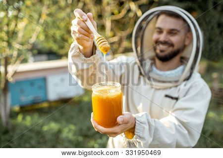 Portrait Of A Handsome Beekeper In Protective Uniform Standing With Honey In The Jar, Tasting Fresh 