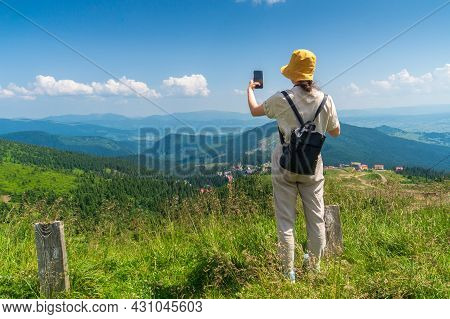 Traveler In A Hat, Resting In Nature. A Tourist Girl Photographs A Panorama Of The Mountains By A Mo
