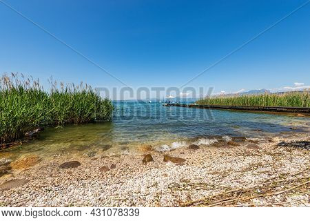 Beach And Wooden Pier With Beach Umbrella And Empty Deck Chairs On Lake Garda, Between The Small Vil