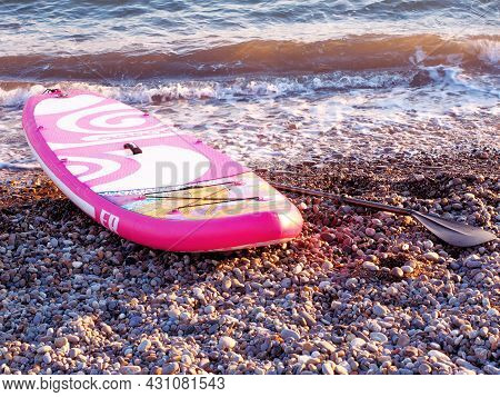 Russia, Dagomys 11.06.2021. Pink Patterned Stand Up Paddle Board On A Pebble Beach Near The Sea In S