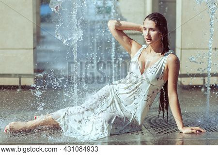 Wet Woman Sits In A Fountain. Her Dress And Hair Are Wet. The Fountain Pours Over The Woman.
