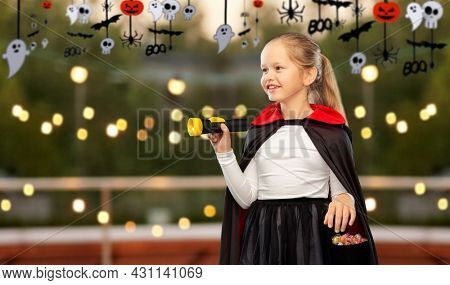 halloween, holiday and trick-or-treating concept - happy smiling girl in black dracula cape or costume with candies and flashlight over roof top party and decorations on background