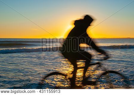 Blurred Image Of Cyclist Riding On Papamoa Beach Back Lit By Sunrise On Distant Horizon