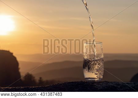 A Glass Of Pure Transparent Mineral Drinking Water Against The Background Of A Sunset In The Mountai