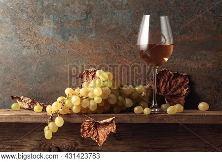 Glass Of White Wine With Grapes And Dried Up Vine Leaves. Old Wooden Table And Vintage Background.