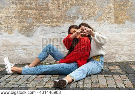 Two Best Friends Hugged Sitting On A Paved Sidewalk By An Old Wall