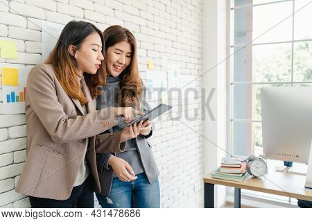 Two Asian Young Creative Businesswomen Holding A Cup Of Coffee, Working On Laptop And Disscuss About