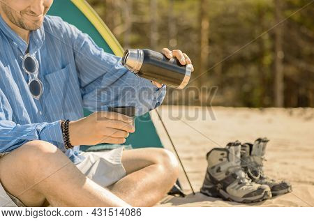 Smiling Caucasian Man Is Sitting Next To His Tourist Tent And Holding A Thermos Bottle Of Tea In His