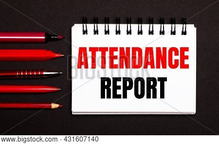 A White Notebook With The Text Attendance Report Next To Red Pens, Pencils And Markers On A Black Ba