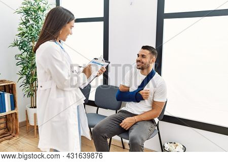 Youg latin injuried man wearing sling talking with the doctor at clinic waiting room.