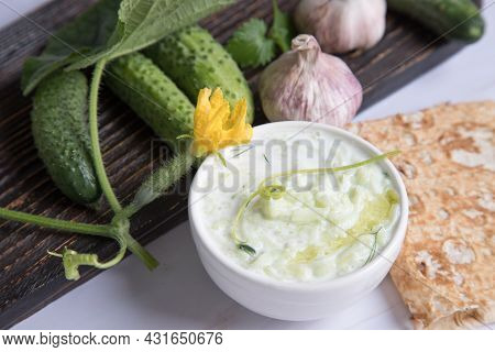A Deep White Plate With Tzatziki Sauce With Pita Bread And Cucumbers On A Wooden Board In The Backgr