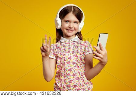 Adorable Kid Shows Peace Gesture, Uses Smartphone, Listens Favorite Music Via White Headphones Wears
