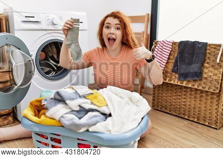 Young redhead woman putting dirty laundry into washing machine doing happy thumbs up gesture with hand. approving expression looking at the camera showing success. 