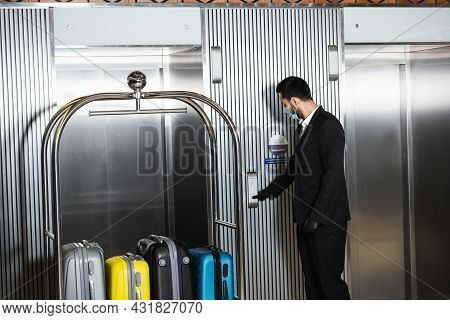 Bellboy In Medical Mask Standing Near Bell Cart Near Elevator In Hotel