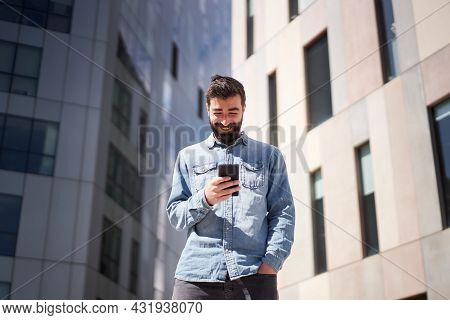 Young Businessman In Denim Shirt Using Smart Phone Near Office Buildings In City Financial Center.