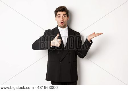 Portrait Of Hadnsome Bearded Man In Formal Suit, Showing Thumb-up And Holding Product In Hand Over W