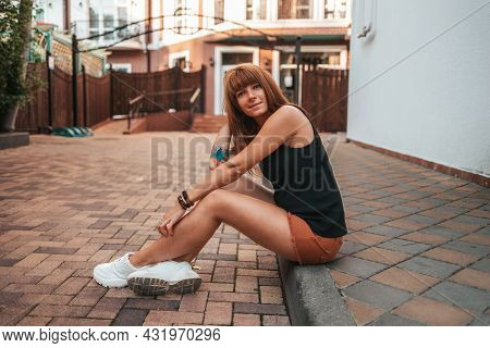 Pretty Smiling Young Woman With Tattooes Is Sitting On The Sidewalk. Side View. The Concept Of Relax
