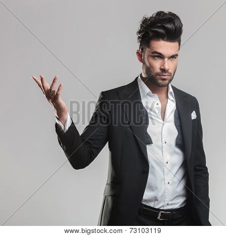 Close up of an elegant young man in tuxedo holding one hand in the air while looking away from the camera. On grey background.