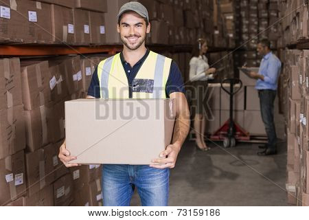 Warehouse worker smiling at camera carrying a box in a large warehouse