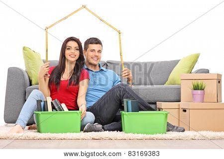 Young couple sitting by a bunch of moving boxes and making a roof over their heads with a tape measure isolated on white background