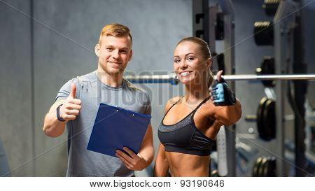 fitness, sport, exercising and diet concept - smiling young woman and personal trainer with clipboard showing thumbs up in gym