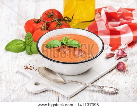 Cold gazpacho soup in bowl with ice, hot pepper and basil served with fresh tomotoes on ceramic board over white wooden background, selective focus, horizontal composition