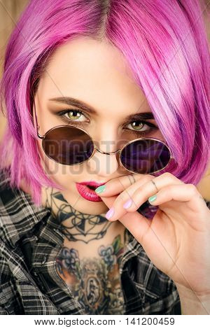Close-up portrait of a modern girl with bright crimson hair and round sunglasses. Tattoo. Hair coloring. Optics style.