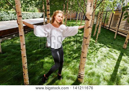 Portrait of young dreamy blonde woman posing among birches on sunny summer day