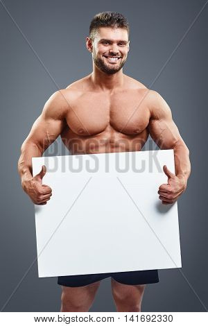 Smiling naked muscular man covering with a white banner isolated over grey background. Vertical portrait of fitness man holding a white paper and showing thumbs up with copy space