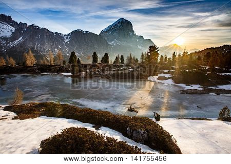 Winter mountain landscape with frozen lake in the front and dramatic sky of evening sun in the background sunrays protruding trees. Wilderness raw nature concept and background. Limides Dolomites.