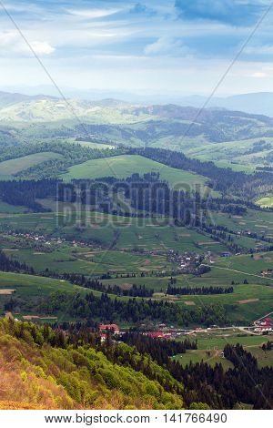 landscape consisting of a Carpathians mountains with fir-trees and green trees and grassy green valley with little houses and blue cloudly sky