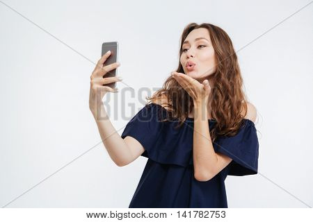 Happy cute young woman sending a kiss and taking selfie with smatphone over white background