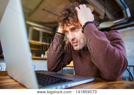 Stressful upset desperate handsome curly man in brown sweetshirt working using laptop and having headache