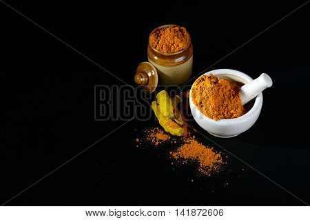 Turmeric powder in mortar with pestle and roots with clay pot on black background