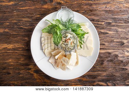 Cheese plate on a dark wooden table. Many kinds of cheese with basil parsley dill and pita on a plate. Top view