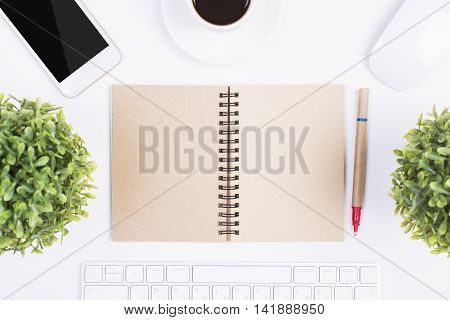 Close up of blank spiral notepad pen plants computer keyboard mouse smartphone and coffee cup on white office desktop. Top view Mock up