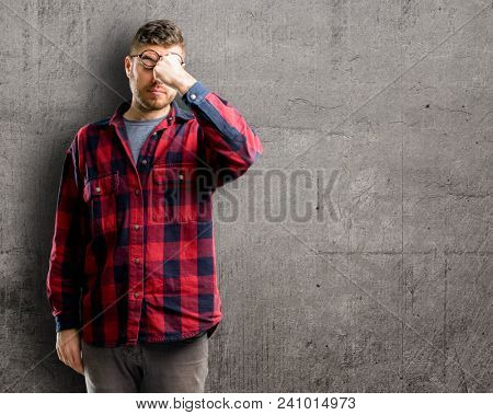 Young handsome man with sleepy expression, being overworked and tired, rubbes nose because of weariness