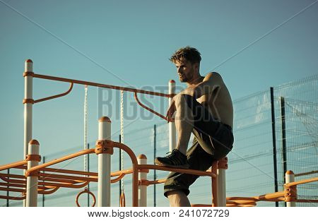 Sportsman With Bare Torso On Monkey Bars Top. Man Sit On Metal Jungle Gym On Blue Sky. Climb, Climbi