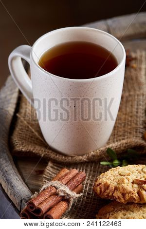 White Porcelain Cup Of Hot Tea, Cookies, Cinnamon Sticks Bunch On A Wooden Barrel Over Rustic Backgr