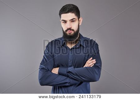 Young Handsome Man Leaning Against Grey Wall With Arms Crossed. A Serious Young Man With A Beard Loo