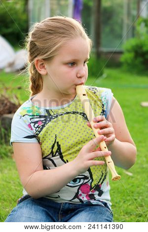 Portrait Of Beautiful Little Girl With Blond Hair In Jeans Playing Flute In The Garden Sitting On Th