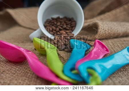 Loose Roasted Coffee Beans, White Porcelain Coffee Mug And Color Plastic Dose Measuring Spoons On Ju