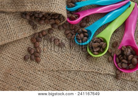 Loose Roasted Coffee Beans And Color Plastic Dose Measuring Spoons On Jute Sack. Conceptual Of Recom
