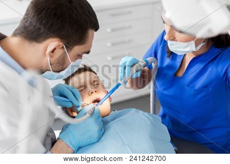 medicine, dentistry and healthcare concept - dentist and assistant with dental drill and saliva ejector treating kid patient teeth at dental clinic