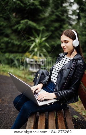 Smiling Student Sitting On Bench Listening Music And Holding Laptop In Park At University Preparing